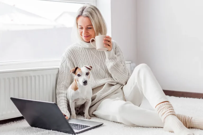 Happy middle aged woman with dog using her laptop at cozy white home thinking about affordable insulation solutions
