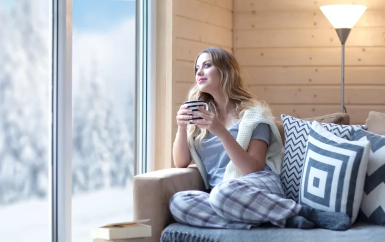 Young woman with cup of coffee sitting home in living room by the window Winter snow landscape view Lazy day off concept while experts are in the attic insulating Efficient Attic Insulation