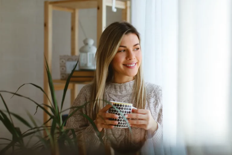 Young woman looking out the window and holding a cup of coffee feeling happy about her Budget-Friendly Insulation