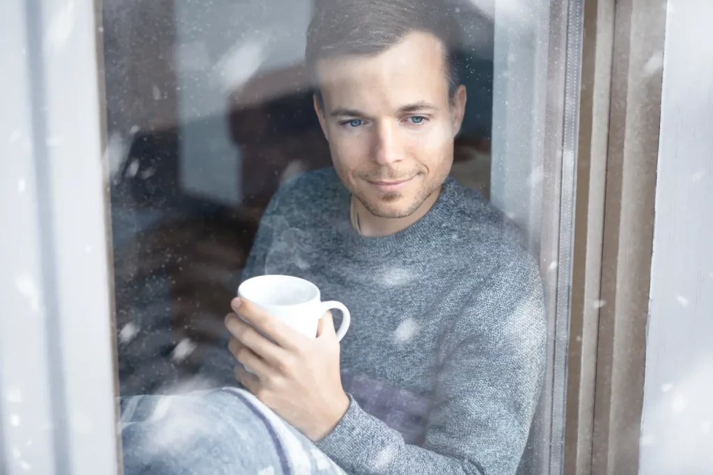 A man sitting on a window ledge, holding a coffee mug. He is looking out a window at the snow.